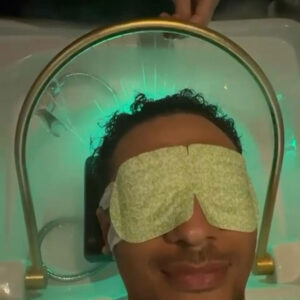 A mans head is under a Japanese Head Spa machine and he is wearing a green eye mask.